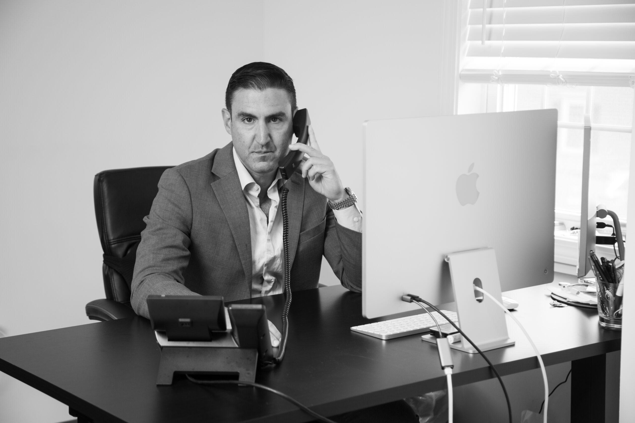 a man in a suit on the phone at his desk and looking at the camera
